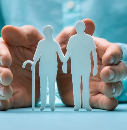 Hand Protecting Senior Couple Cutout Figures On Desk